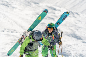 two green suited skiers holding ogo's skis on their backs