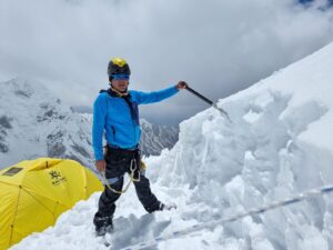 a man shoveling the snow on the mountain with a yellow tent behind him